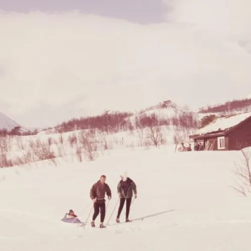 Bilde fra 60-tallet med familie på skitur. Fjell vises i bakgrunnen og to små hytter. I dag er området ved skiheisen utbygget med mange hytter.