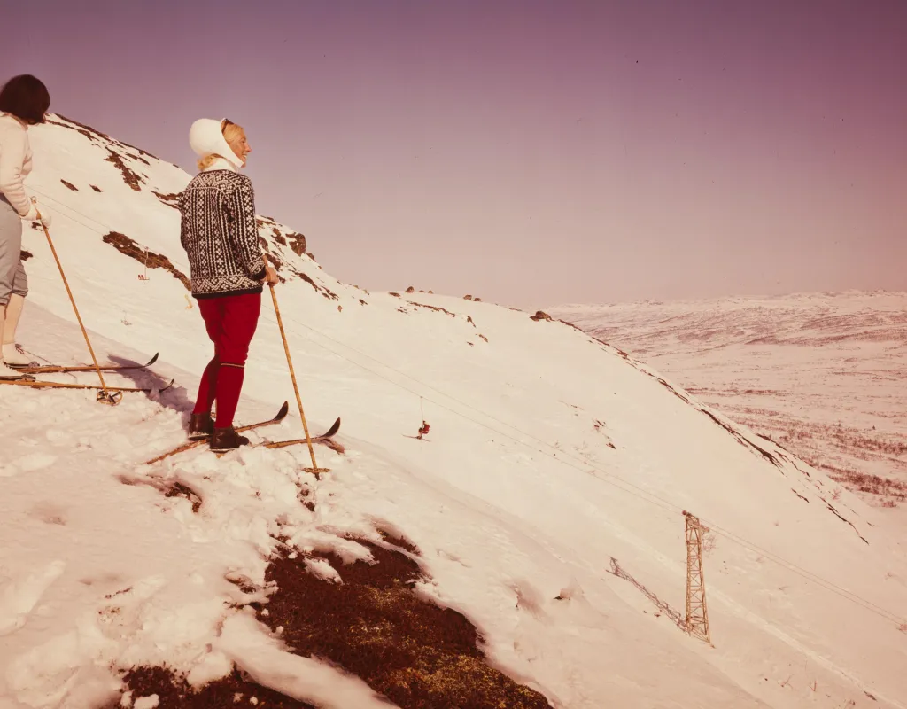 To damer i tidsriktige klær fra 1969 på ski på toppen av Hovden Skisenter. Nyter sol og utsikt.