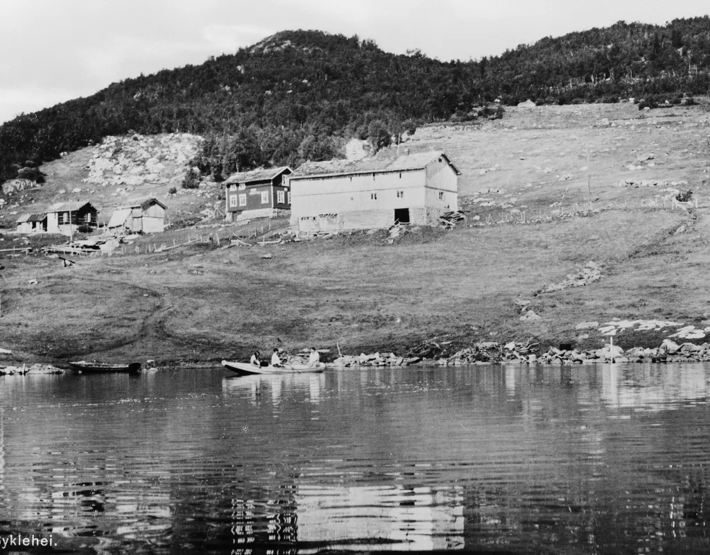 Bjåen Turiststasjon anno 1940. Personer i robåt på vannet med gamle bygninger og kulturlandskap i bakgrunnen.