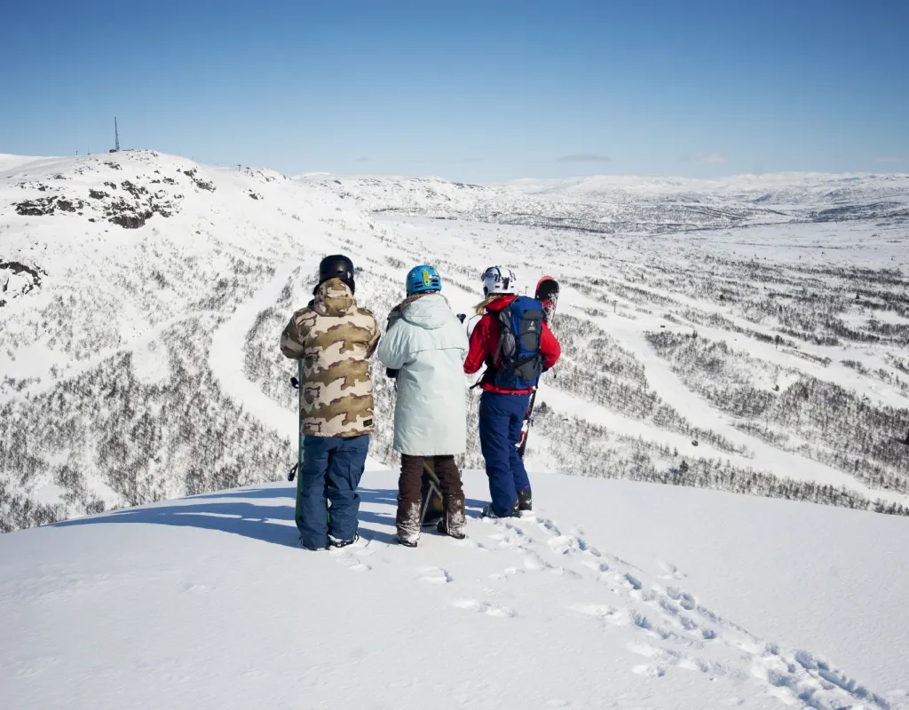 Tre personer med slalomski og snowboard står på Hovdenuten og skuer ut over alle bakkene på Hovden Alpinsenter. 