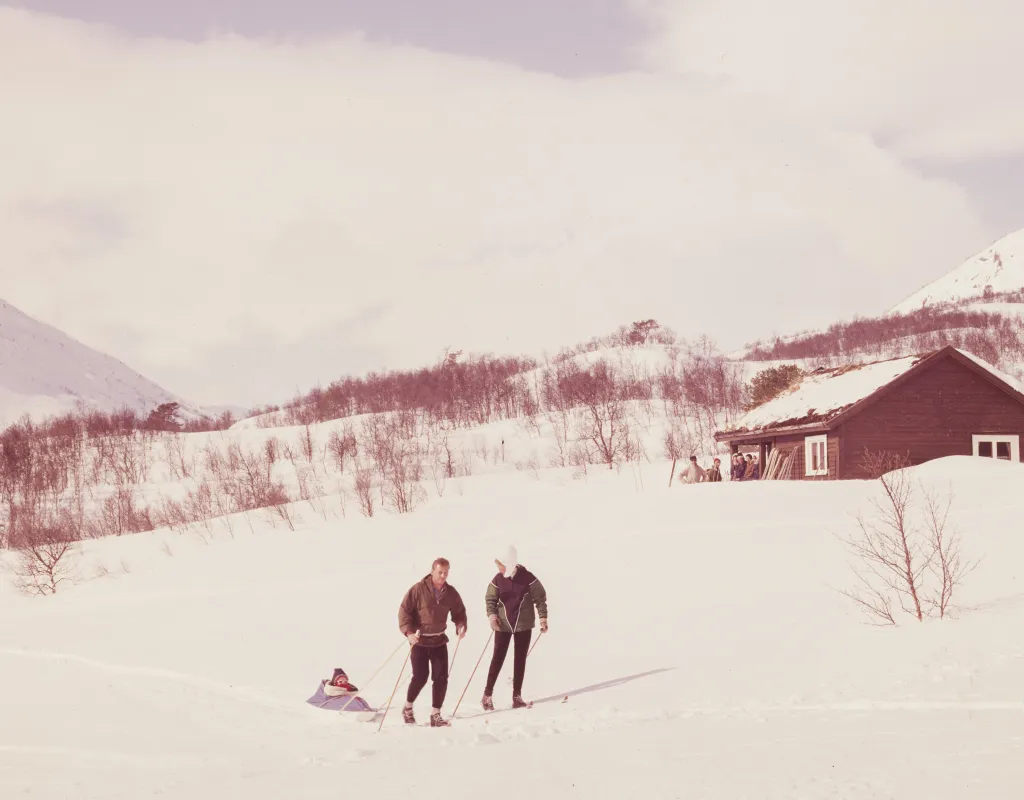 Bilde fra 60-tallet med familie på skitur. Fjell vises i bakgrunnen og to små hytter. I dag er området ved skiheisen utbygget med mange hytter.